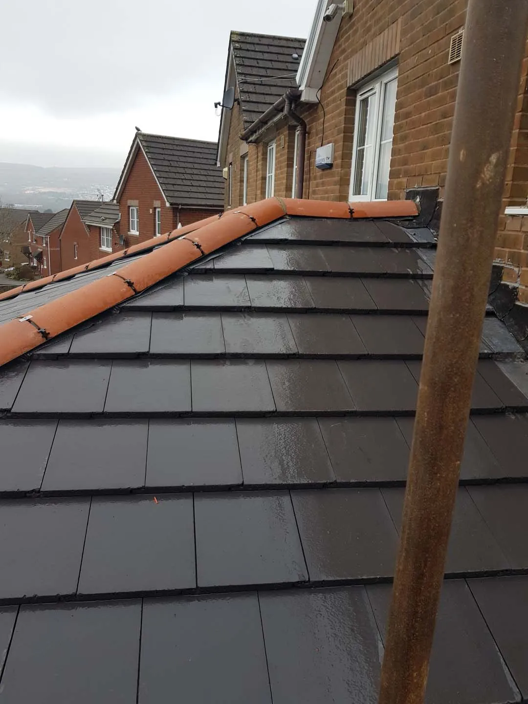 Wet slate-effect tiled roof with clipped clay hip tiles meeting a brick house wall