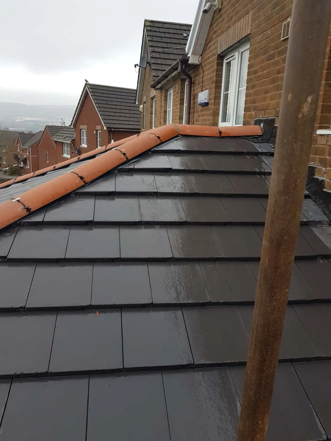 Wet slate-effect tiled roof with clipped clay hip tiles meeting a brick house wall