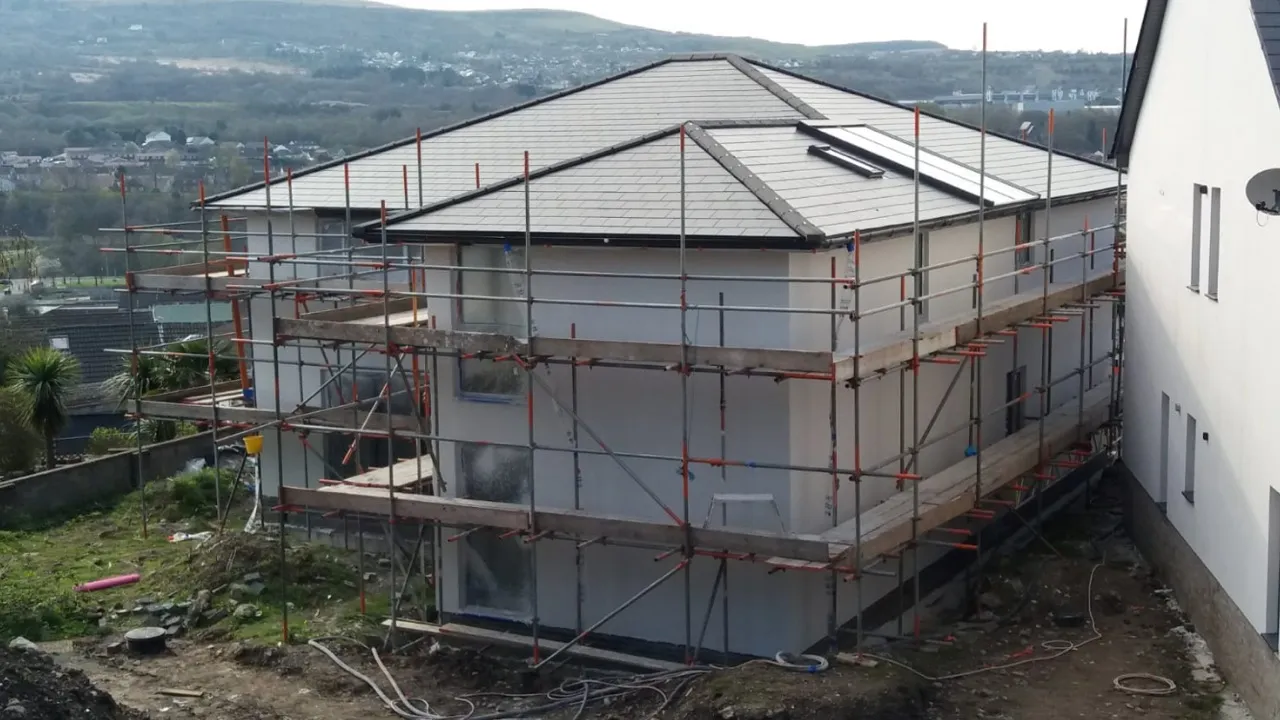 Two-storey new-build house with a hipped slate-effect tiled roof and rooflights, surrounded by scaffolding